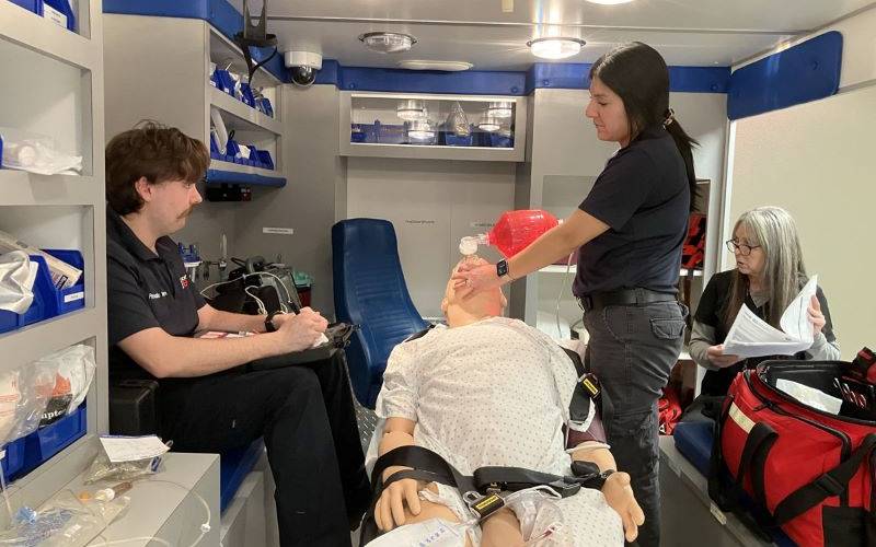 two medical students in the back of a staged ambulance to practice medical procedures on a dummy in a stretcher